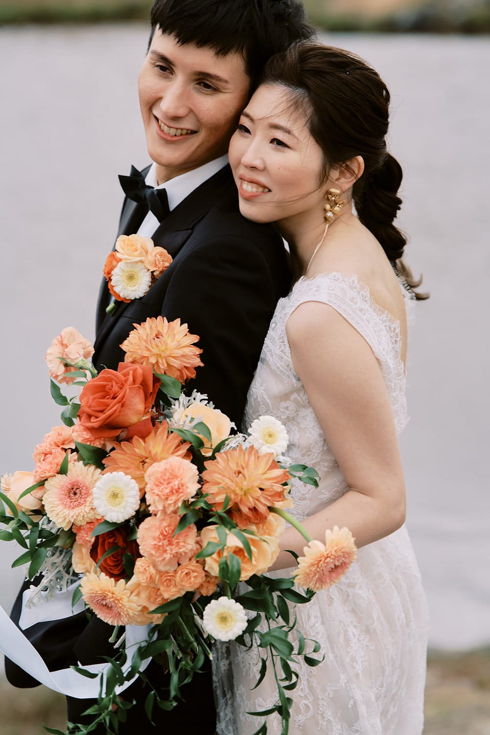 Queenstown New Zealand Heli Wedding Elopement Photographer クイーンズタウン　ニュージーランド　エロープメント 結婚式 | A bride and groom embracing and smiling on their wedding day, with the bride holding a bouquet of orange flowers, captured beautifully by YURI, a Queenstown Wedding Photographer.